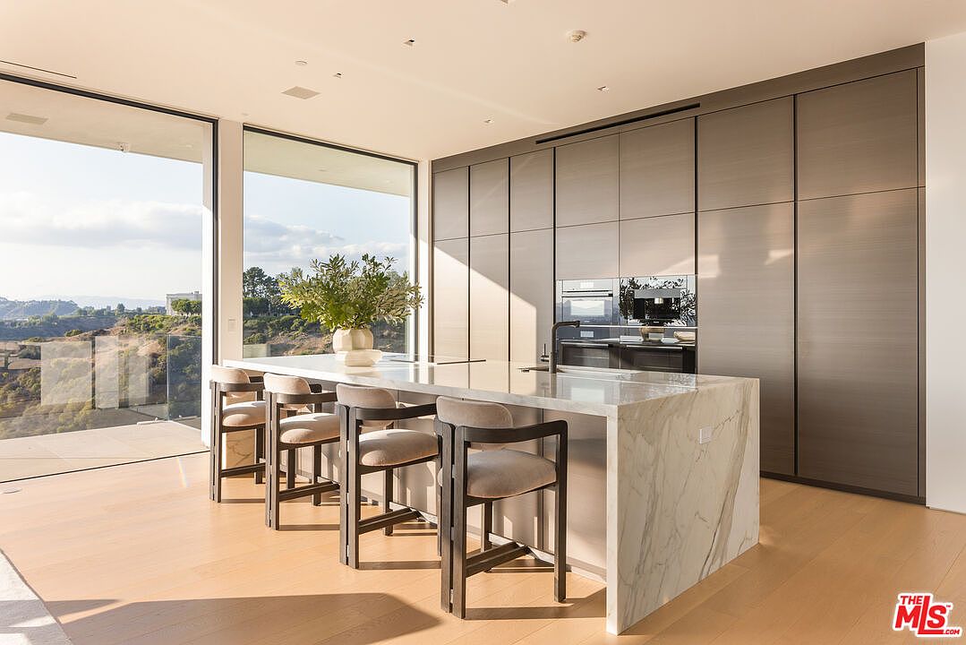 This is a modern kitchen featuring a large marble island with seating, sleek gray cabinetry, and high-end appliances. The kitchen is well-lit with natural light from large windows that offer a scenic view. The overall design is minimalist and luxurious, creating a sophisticated and inviting space.
