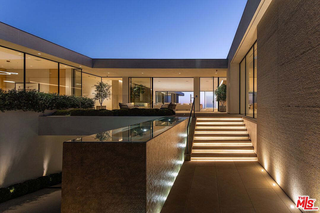 This image showcases a modern home's entryway at dusk, featuring illuminated stairs leading up to a sleek, glass-walled structure. A reflecting pool adds a luxurious touch, while the minimalist landscaping and warm lighting create an inviting and sophisticated ambiance. The exterior is characterized by clean lines and a neutral color palette.