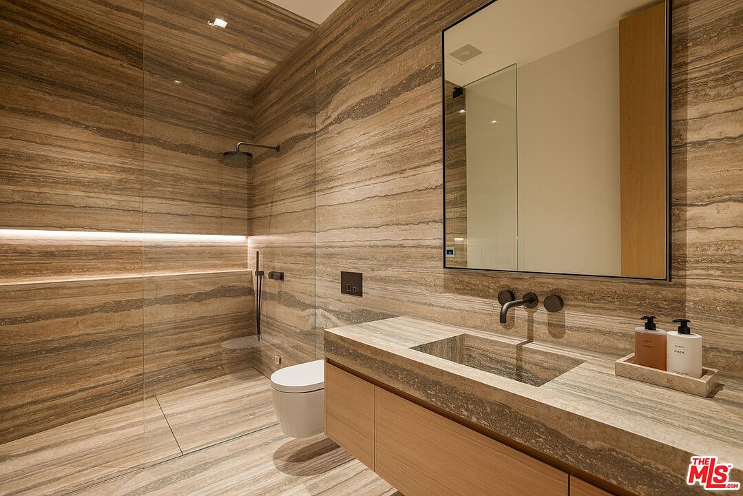 This is a modern primary bathroom featuring horizontally-laid travertine walls and flooring, creating a luxurious and spa-like atmosphere. The bathroom includes a floating vanity with a stone countertop and integrated sink, a large mirror, and a glass-enclosed shower with a rainfall showerhead. The overall design is sleek and minimalist, emphasizing natural materials and clean lines.