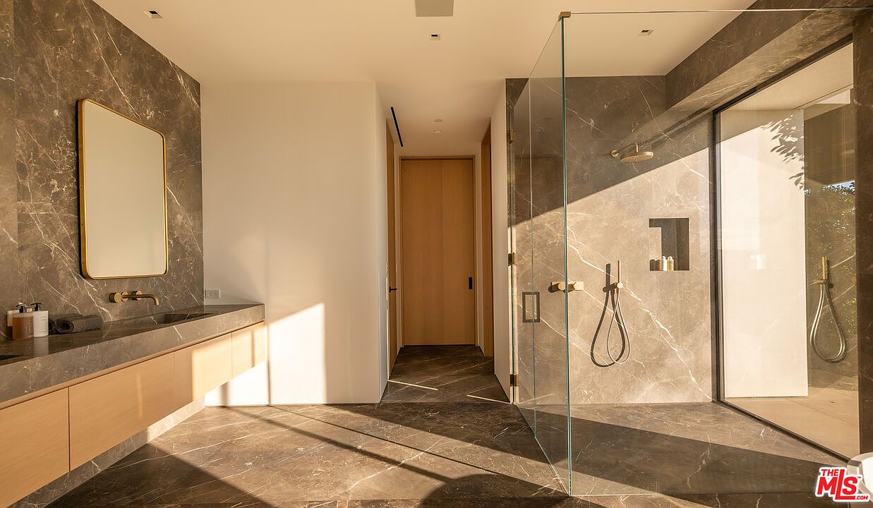 This is a modern primary bathroom featuring marble walls and flooring, a floating vanity with a rectangular mirror, and a glass-enclosed shower. The shower has both a rainfall showerhead and a handheld sprayer. The design is sleek and luxurious, with a focus on natural materials and clean lines.