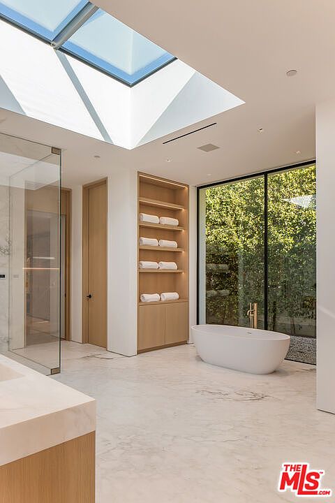 This is a bright and modern primary bathroom featuring a freestanding white bathtub positioned in front of a large window overlooking lush greenery. The room is illuminated by a large skylight, and built-in shelving provides storage for towels. The flooring is a light-colored marble, contributing to the bathroom's luxurious and spa-like atmosphere.