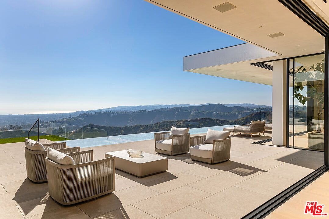 This image showcases a luxurious outdoor patio area with modern furniture, including armchairs and a coffee table, arranged on a tiled deck. The patio overlooks a stunning view of rolling hills and a distant coastline, with a sleek infinity pool adding to the upscale ambiance. Large sliding glass doors seamlessly connect the indoor and outdoor spaces, highlighting the property's contemporary design and emphasis on indoor-outdoor living.