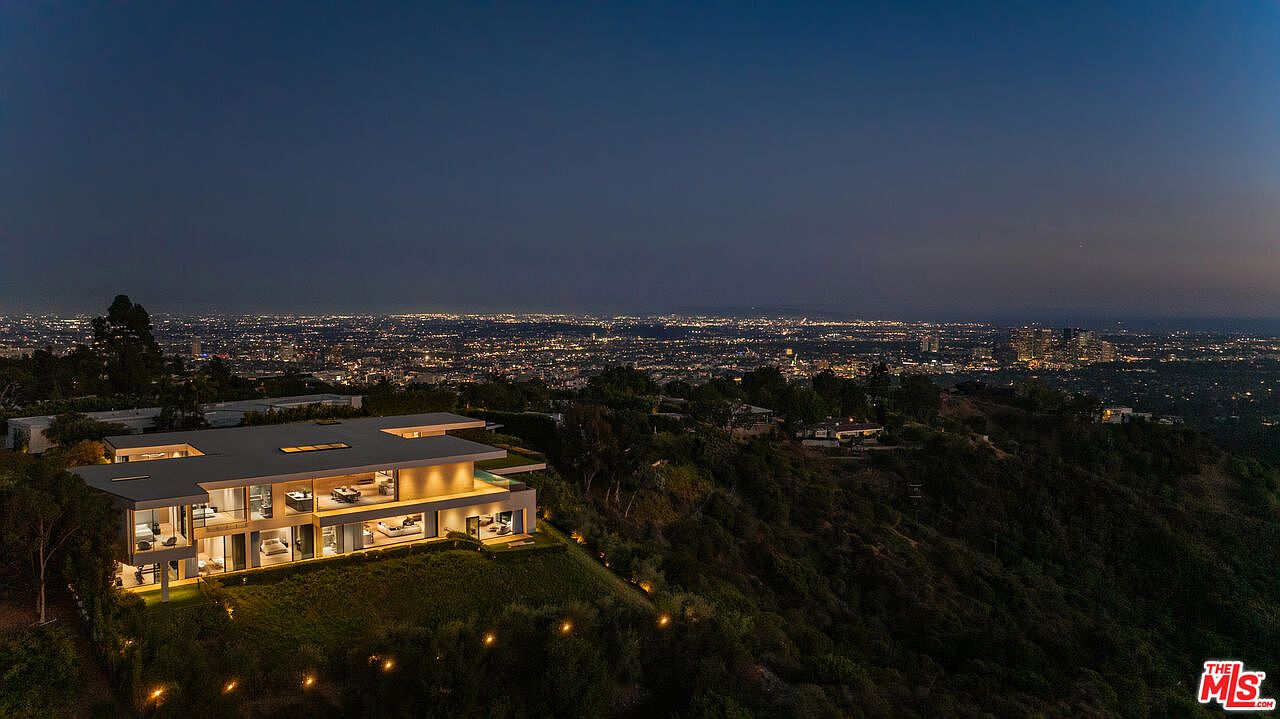 This aerial view showcases a modern, multi-level home perched on a hillside, offering stunning panoramic city views at night. The architecture features clean lines, large glass windows, and illuminated interiors, creating a luxurious and inviting ambiance. The surrounding landscape is lush and green, adding to the property's exclusivity and appeal.