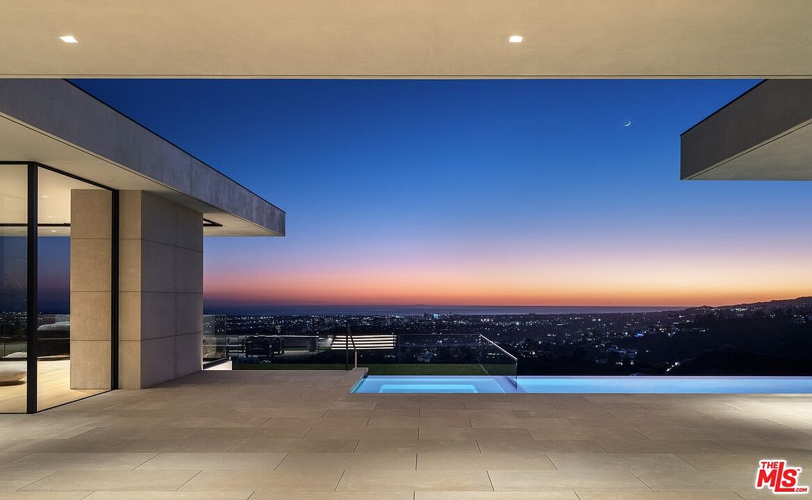 This image showcases a luxurious outdoor space featuring a modern infinity pool with a glass railing, offering stunning panoramic views of the city and the horizon at dusk. The pool is integrated into a spacious tiled patio area, part of a contemporary architectural design with clean lines and minimalist aesthetics. The scene evokes a sense of tranquility and high-end living.
