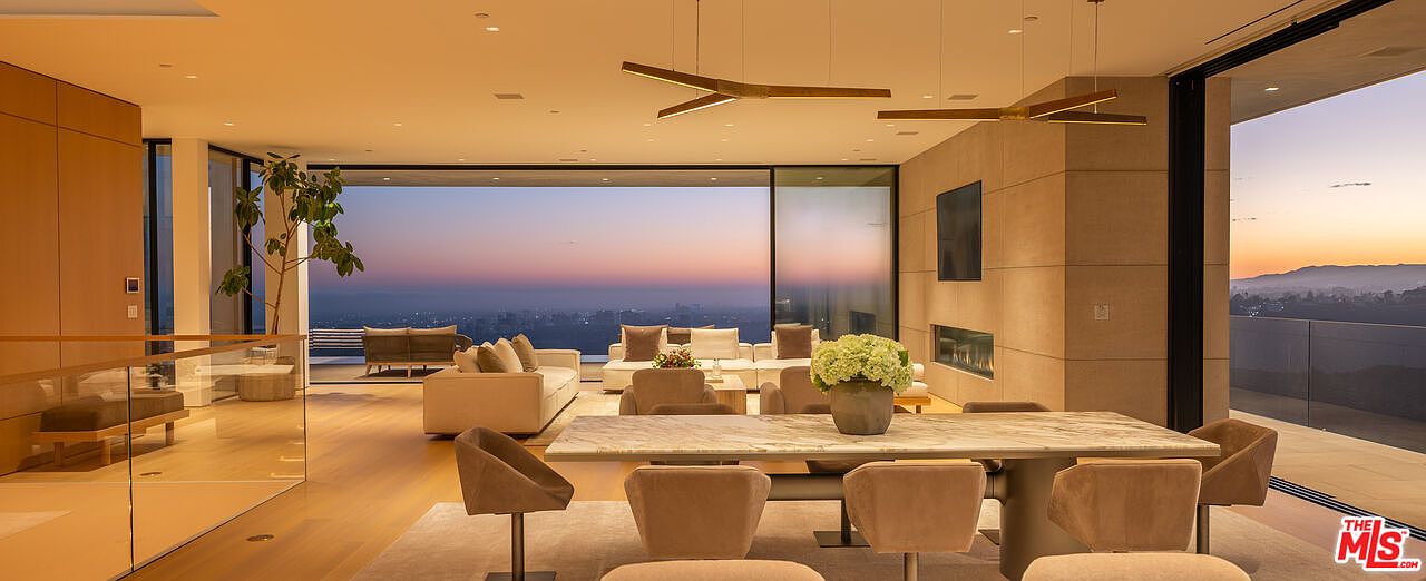 This is a luxurious living and dining area featuring floor-to-ceiling windows that offer a stunning panoramic view of the city at sunset. The space is furnished with modern sofas, chairs, and a marble dining table, all set on a plush rug. The room is illuminated by contemporary pendant lighting, creating a warm and inviting atmosphere.