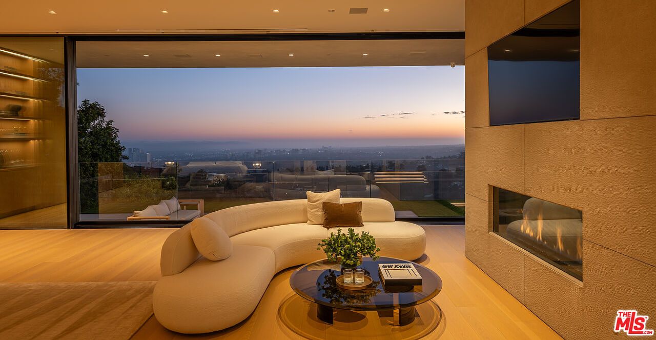 This is an interior shot of a luxurious living room featuring a curved white sofa, a glass coffee table with books and plants, and a modern fireplace. Large glass doors offer a stunning view of the city skyline at dusk, creating a seamless indoor-outdoor living experience. The room is well-lit with natural light and features hardwood floors, enhancing the elegant and contemporary design.
