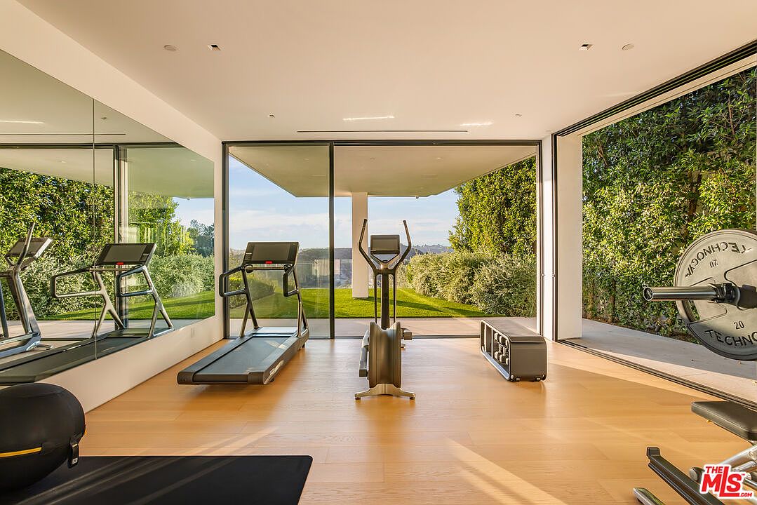 This is a well-lit home gym featuring modern exercise equipment such as treadmills, an elliptical machine, and weightlifting equipment. The room has large windows offering views of the outdoors, and a mirrored wall enhances the sense of space. The hardwood floors and minimalist design create a clean and inviting workout environment.