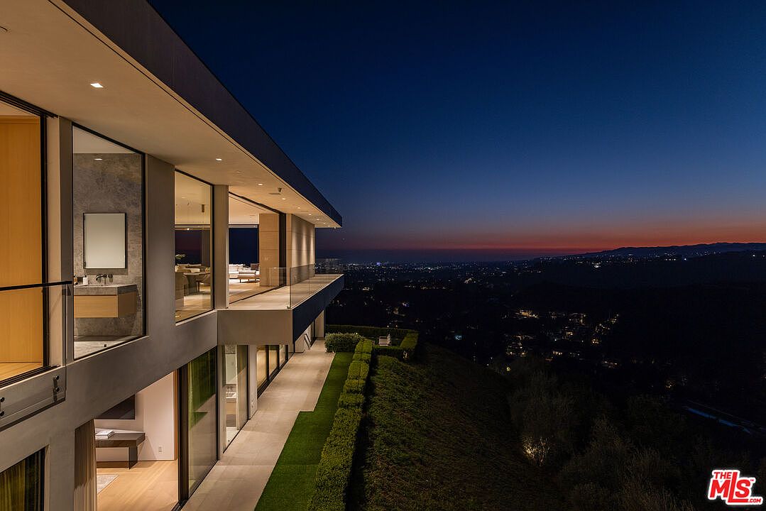 This image showcases the rear exterior of a modern luxury home at dusk, highlighting its sleek architectural design and expansive outdoor living spaces. The multi-level structure features large glass windows and doors that offer panoramic views of the city skyline and surrounding landscape. A well-manicured lawn and minimalist landscaping add to the property's sophisticated appeal, creating an inviting atmosphere for relaxation and entertainment.