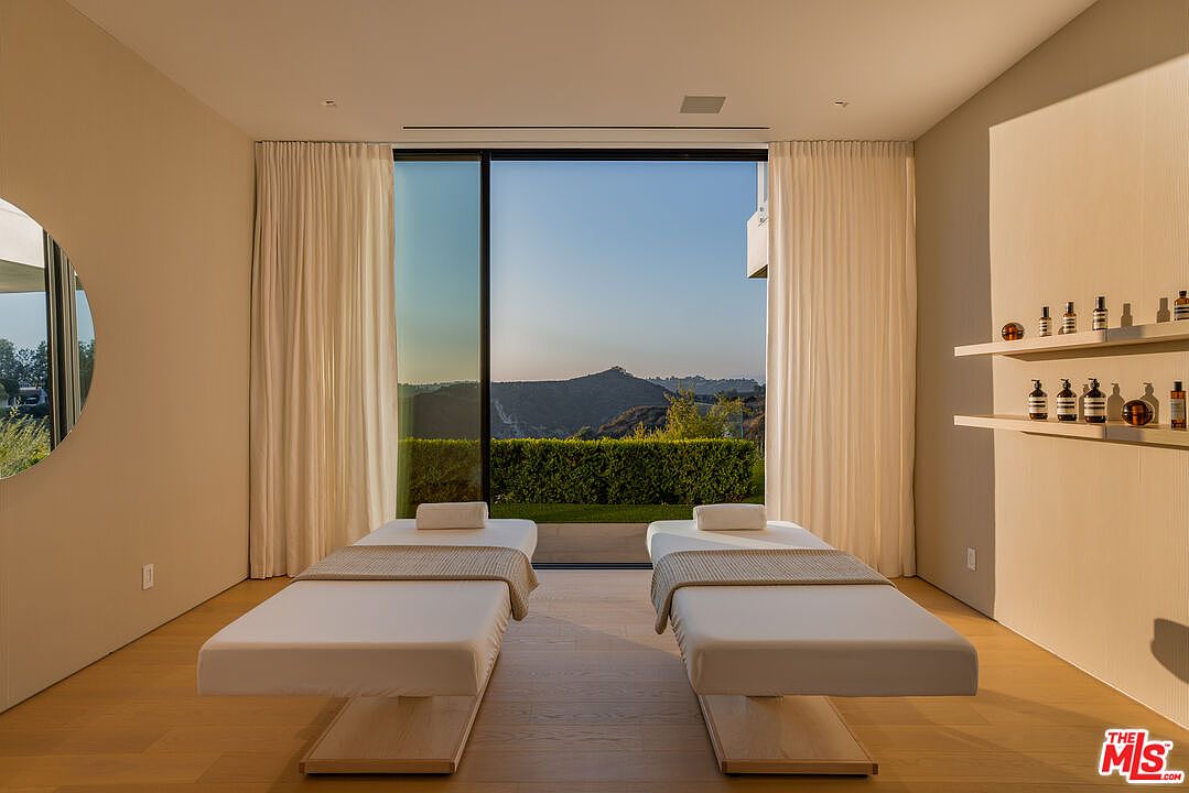 This interior shot showcases a serene spa room featuring two massage tables positioned in front of a large window with a scenic mountain view. The room is decorated in neutral tones with light wood flooring, cream-colored curtains, and minimalist shelving displaying spa products. The overall impression is one of tranquility and luxury.