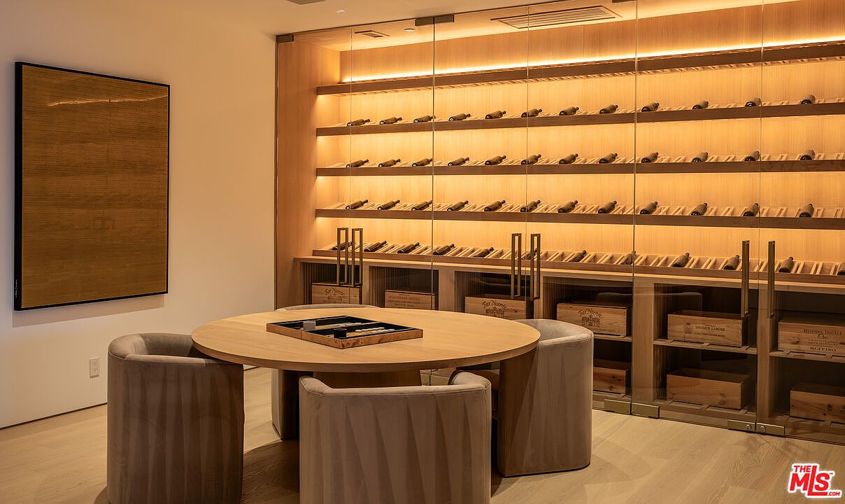 This image showcases a sophisticated wine cellar featuring a well-lit, temperature-controlled environment. The cellar is equipped with wooden shelving displaying numerous wine bottles, complemented by wooden storage boxes below. A round wooden table with four upholstered chairs creates a tasting area, while a modern art piece adorns the adjacent wall, adding a touch of elegance.