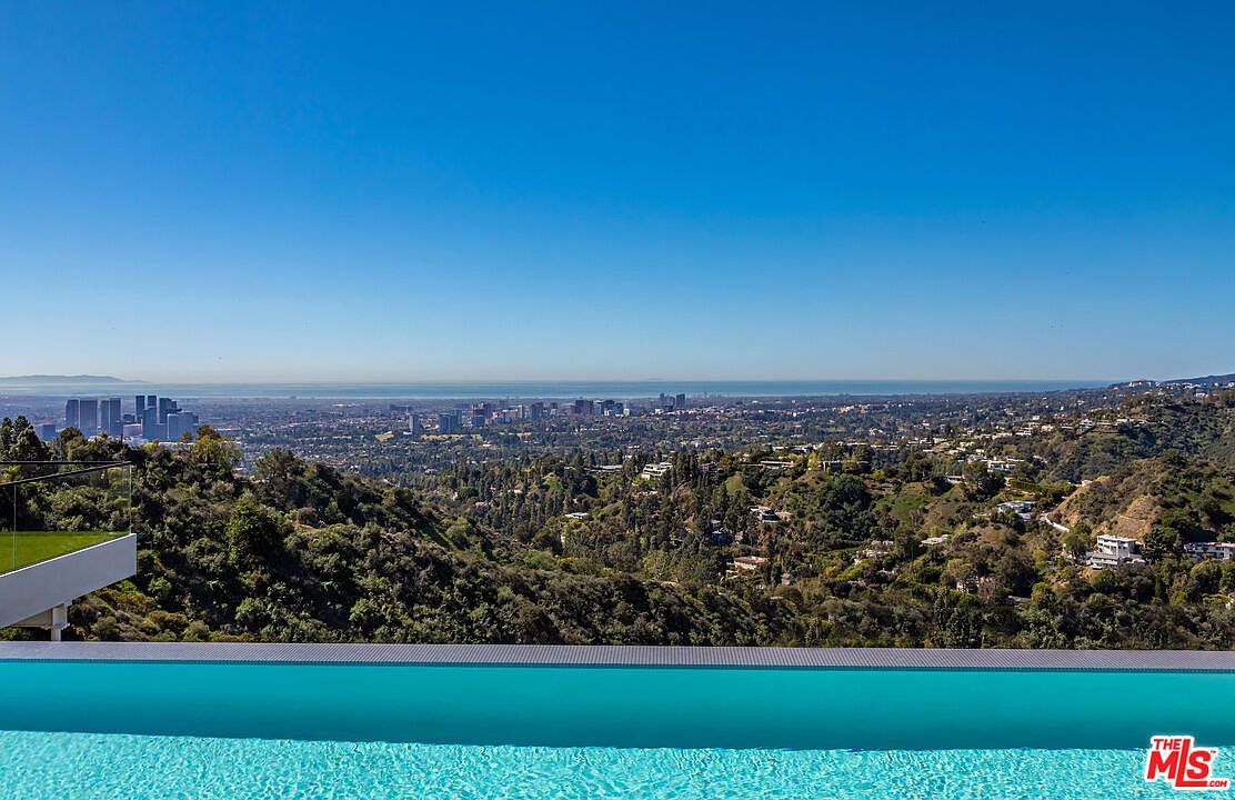 This image showcases a stunning infinity pool overlooking a vast cityscape and lush hillside. The pool's turquoise water contrasts beautifully with the clear blue sky, creating a sense of luxury and tranquility. The view emphasizes the property's prime location and desirable outdoor living space.