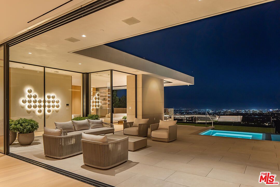 This image showcases a luxurious outdoor patio area with modern furnishings, including a sofa and armchairs, set against a backdrop of a stunning city view at night. The patio seamlessly connects to the interior through large glass doors, and a sleek pool adds to the upscale ambiance. The architectural design emphasizes clean lines and a sophisticated aesthetic, perfect for entertaining or relaxing.