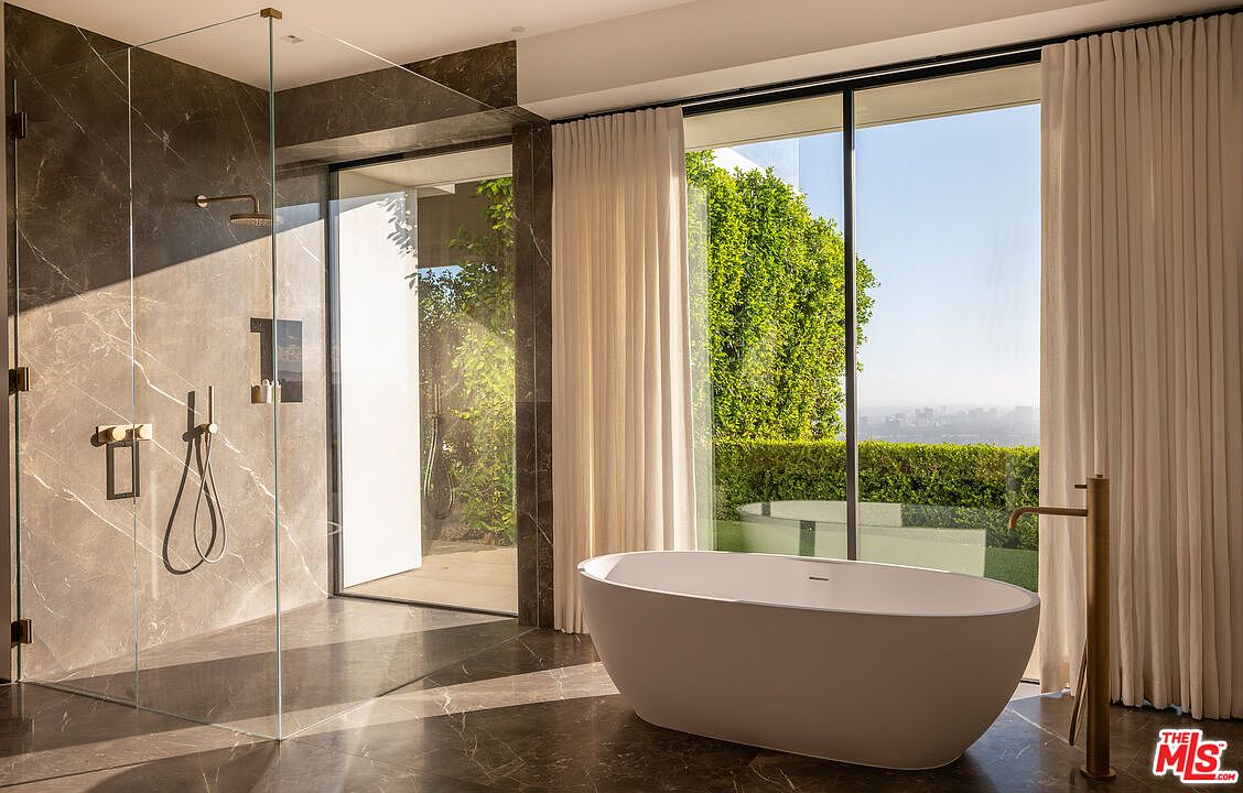 This is a luxurious primary bathroom featuring a modern freestanding bathtub positioned in front of a large window with a view of lush greenery. The bathroom also includes a glass-enclosed shower with elegant fixtures and marble-like wall panels, creating a spa-like atmosphere. The dark flooring complements the light-colored tub and curtains, enhancing the room's sophisticated and serene ambiance.