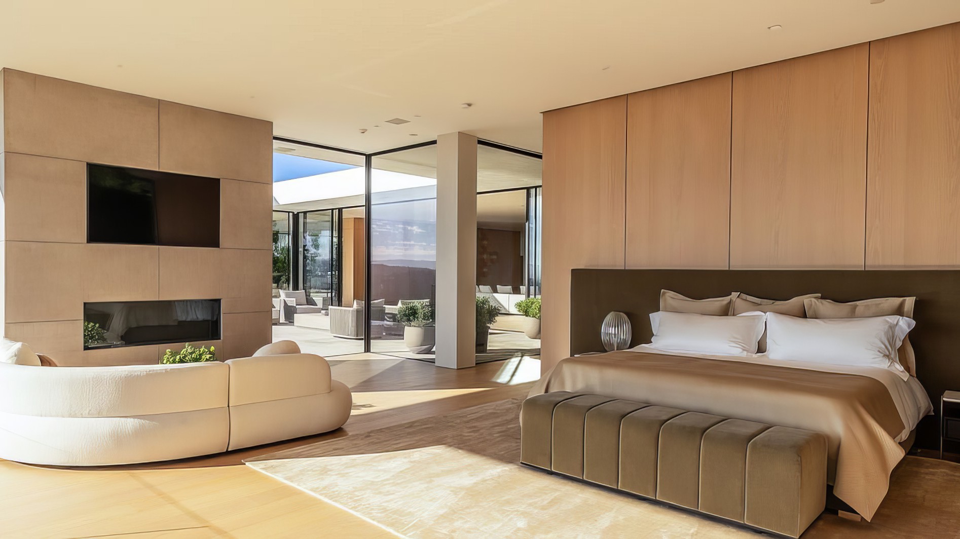This is a spacious primary bedroom featuring a modern design with natural wood paneling. The room includes a king-size bed with neutral-toned bedding and a minimalist aesthetic. Large glass doors offer access to an outdoor patio, creating an seamless indoor-outdoor living experience.