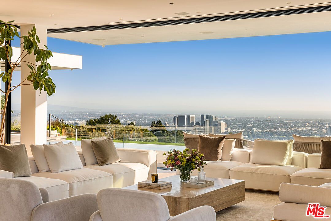 This is a luxurious living room with a panoramic city view. The room features a large, sectional sofa with neutral-toned cushions and throw pillows, a travertine coffee table with decorative items, and modern armchairs. The open floor plan and floor-to-ceiling windows create a bright and airy atmosphere, emphasizing the stunning vista.