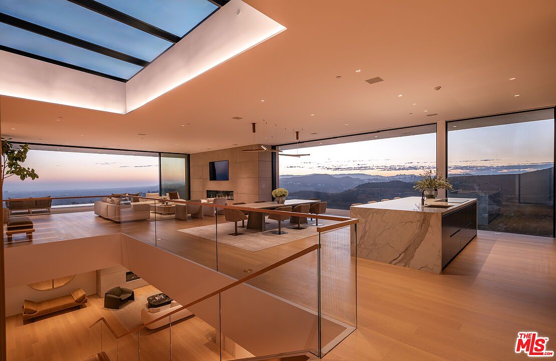 This is a luxurious living room featuring an open floor plan with floor-to-ceiling windows offering panoramic views of the landscape. The space includes a dining area, a kitchen island with marble countertops, and a comfortable seating area. The interior design is modern and sophisticated, with hardwood floors and a neutral color palette, creating a bright and airy atmosphere.