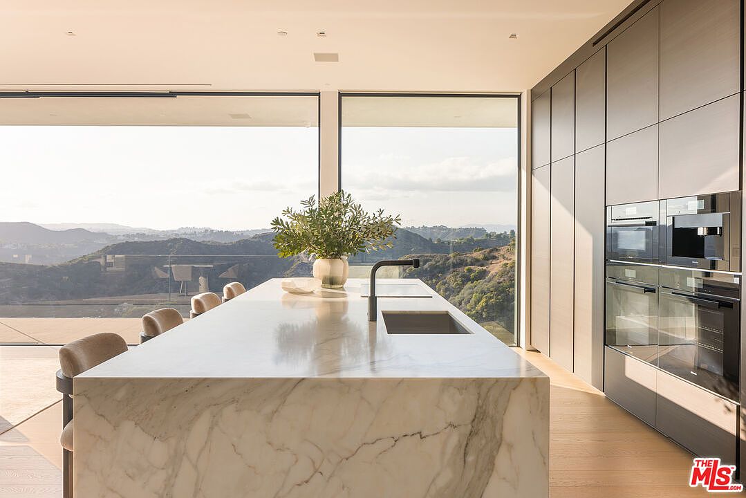 This is a modern kitchen featuring a large marble island with a sink and faucet, surrounded by bar stools. Floor-to-ceiling windows offer expansive views of the surrounding landscape, while sleek, minimalist cabinetry and built-in appliances line the wall. The overall impression is one of luxury and sophistication, emphasizing the seamless integration of indoor and outdoor spaces.