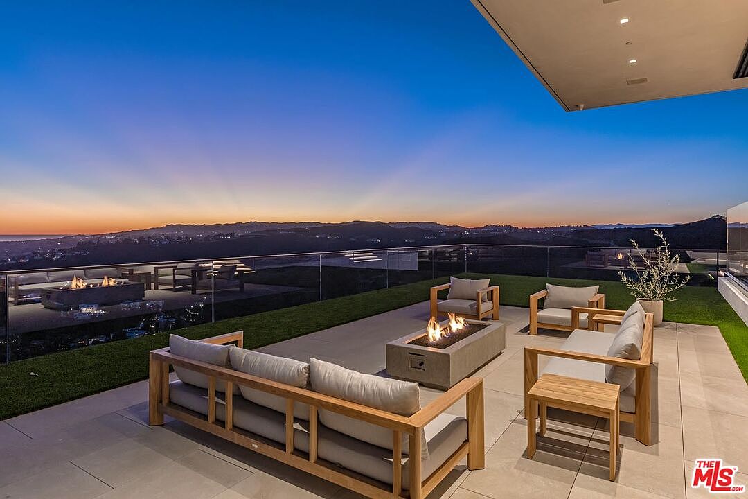 This image showcases a luxurious outdoor patio area at dusk, featuring modern wooden furniture with plush cushions arranged around a concrete fire pit. A glass railing provides unobstructed views of the distant city lights and rolling hills, creating a serene and upscale ambiance. The patio is partially covered with artificial turf, adding a touch of greenery to the space.