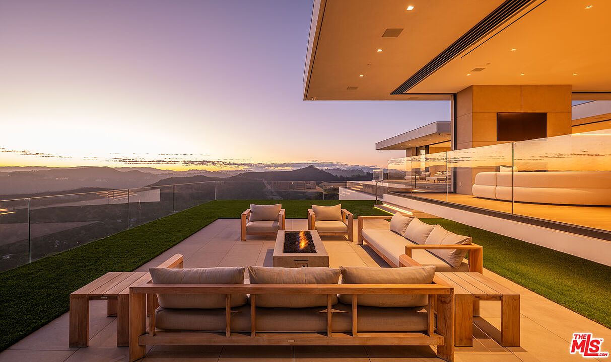 This image showcases a luxurious outdoor patio area with modern furniture, including sofas and chairs arranged around a fire pit. The patio features a sleek design with glass railings, offering stunning panoramic views of the surrounding landscape and a vibrant sunset. The space seamlessly blends indoor and outdoor living, highlighting the property's high-end appeal.