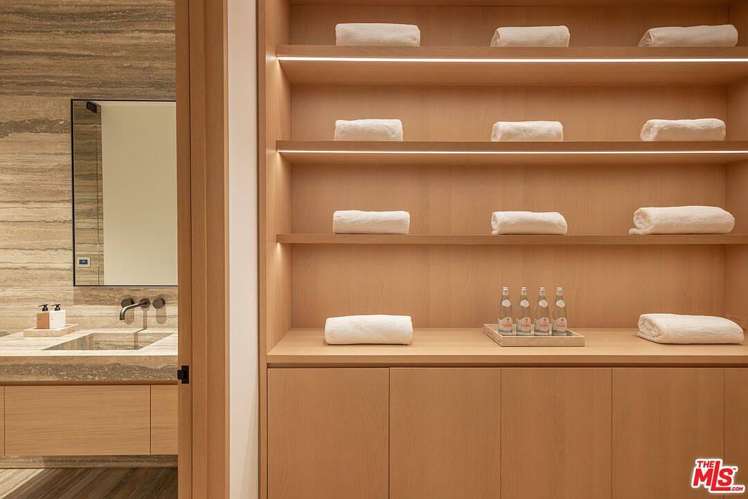 The image showcases a luxurious primary bathroom featuring a modern vanity with a stone countertop and integrated sink. A large mirror hangs above the vanity, reflecting the natural light. To the right, built-in wooden shelves display neatly folded white towels and bottled water, adding a spa-like ambiance to the space.