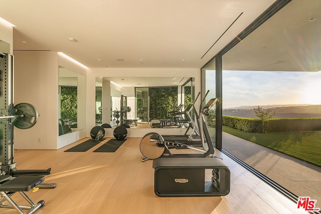 This is a modern home gym featuring state-of-the-art exercise equipment, including treadmills and weight machines. The room has light wood flooring, large mirrors, and floor-to-ceiling windows that offer a stunning view of the landscape. The space is bright, airy, and designed for a premium workout experience.
