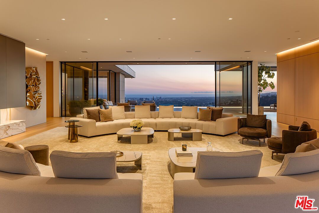 This expansive living room features a neutral color palette with large sectional sofas arranged around multiple coffee tables, creating a comfortable and inviting space. Floor-to-ceiling windows offer panoramic views of the city skyline at dusk, enhancing the luxurious and modern aesthetic. The room is well-lit with recessed lighting, and the open layout promotes a sense of spaciousness.