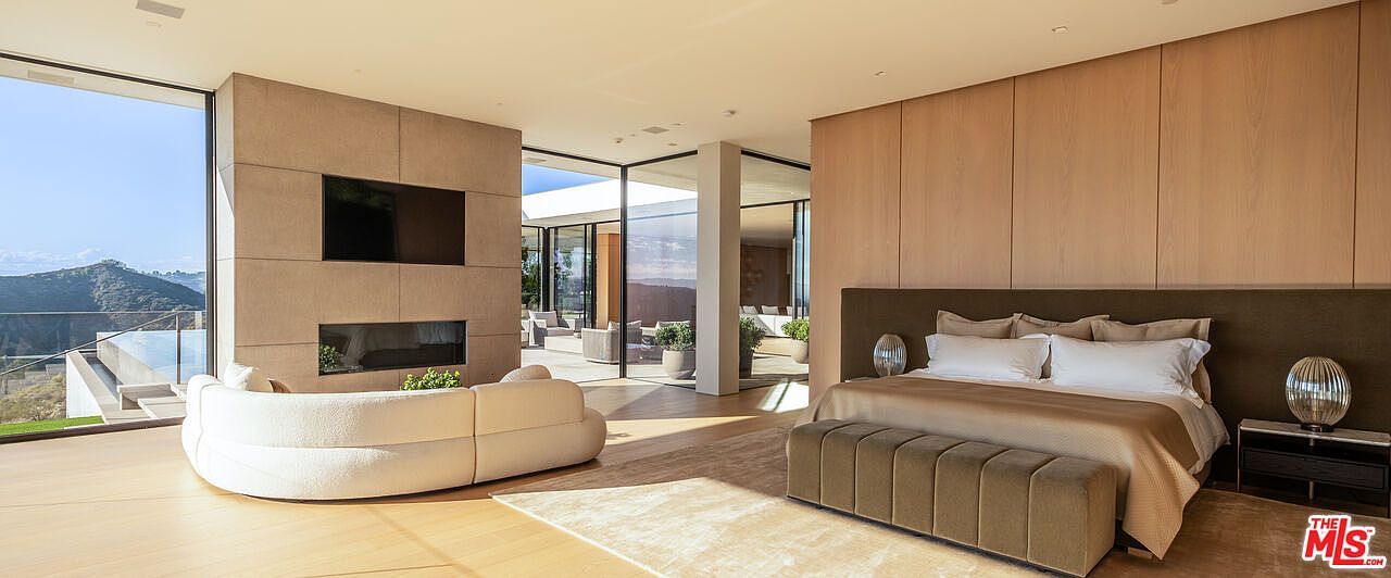 This is a luxurious primary bedroom featuring a modern design with clean lines and natural materials. The room includes a large bed with a padded headboard, a built-in fireplace and television, and floor-to-ceiling windows offering stunning views. The color palette is neutral, creating a serene and inviting atmosphere.