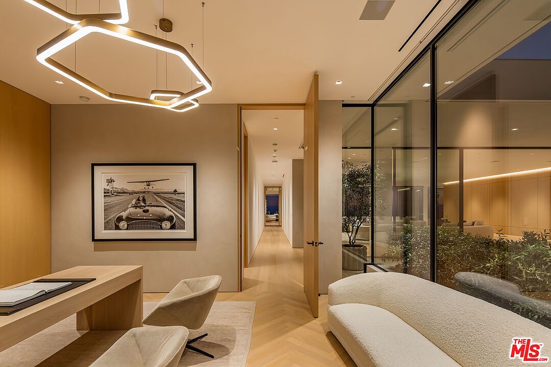 This interior shot showcases a modern office space with a sleek desk, stylish chairs, and a striking black and white photograph on the wall. A unique, geometric light fixture hangs from the ceiling, and large windows offer a view of greenery, creating a bright and inviting workspace. The open doorway leads to a hallway, enhancing the sense of spaciousness.