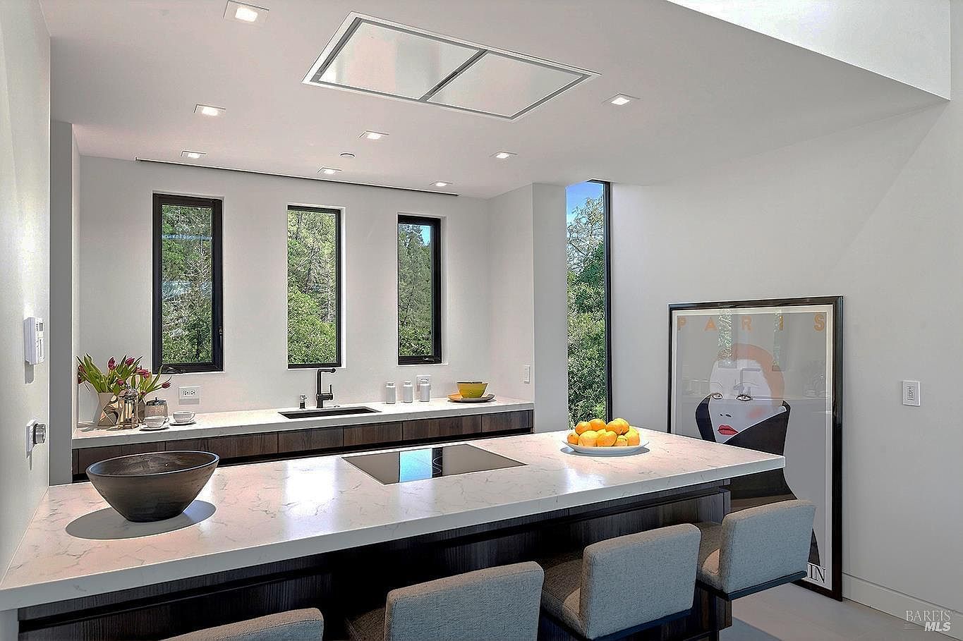 This modern, minimalist kitchen features a large central island with a white marble-patterned countertop and integrated cooktop, complemented by dark wood cabinetry. The space is brightened by recessed lighting and tall, narrow windows that frame a lush green outdoor view, while a large piece of contemporary art adds a bold focal point to the wall. The overall aesthetic is sleek, sophisticated, and highly functional, perfect for a luxury home listing.