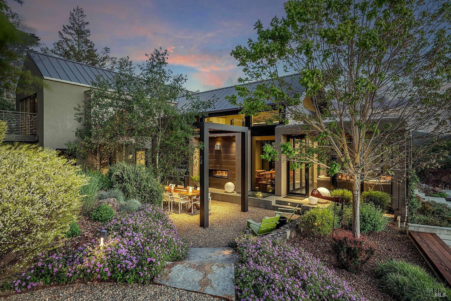 This stunning contemporary home features a seamless blend of indoor-outdoor living, highlighted by a spacious gravel patio with an integrated outdoor fireplace and dining area. The architecture showcases clean lines, large glass walls, and a metal roof, all set against a beautifully landscaped garden with vibrant purple flowers and mature trees. The twilight lighting creates a warm, inviting atmosphere, emphasizing the home's sophisticated and tranquil aesthetic.