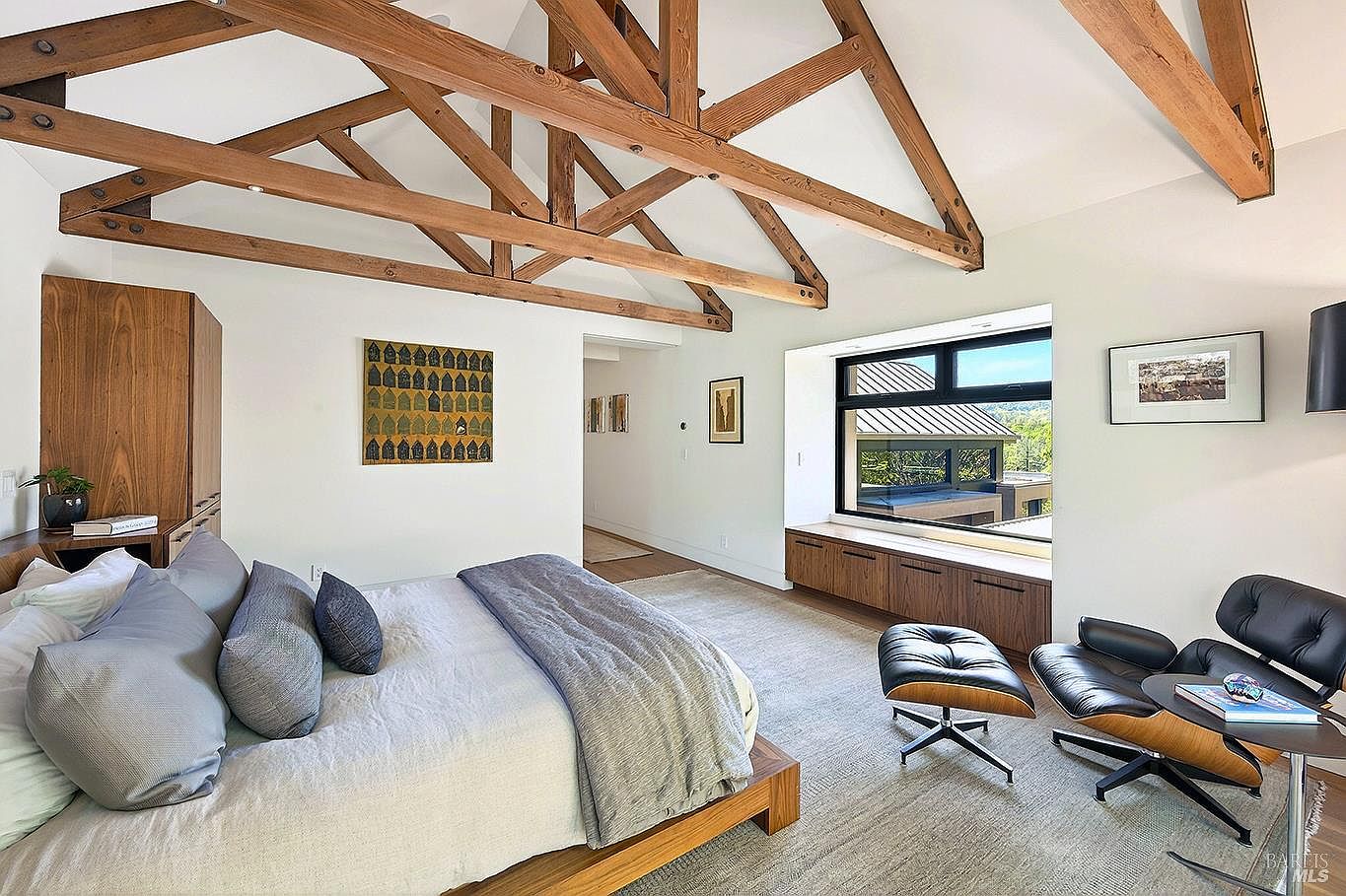 This spacious primary bedroom features a striking vaulted ceiling with exposed wooden trusses, creating a warm, rustic-modern aesthetic. The room is anchored by a platform bed with neutral bedding and complemented by a sleek leather lounge chair and ottoman set near a large window with built-in storage. The clean white walls and soft carpet provide a serene atmosphere, while the large window offers a picturesque view of the surrounding landscape.