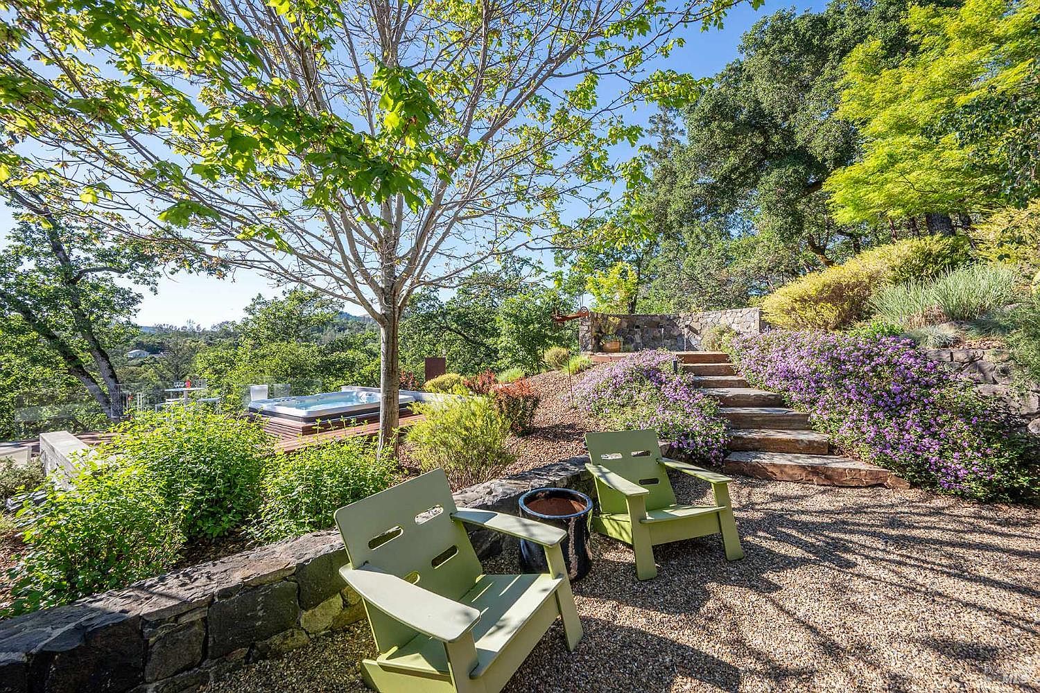 This serene outdoor space features a gravel seating area with two light green Adirondack chairs overlooking a lush, terraced garden. A stone retaining wall frames the foreground, while a hot tub is visible on a wooden deck nestled among mature trees and vibrant purple flowering shrubs. The scene captures a peaceful, elevated perspective of a beautifully landscaped backyard, perfect for relaxation and outdoor entertaining.