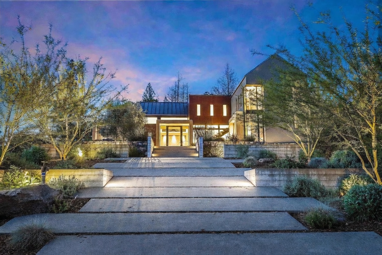 This striking modern residence is captured at twilight, showcasing a dramatic approach via a wide, tiered concrete walkway illuminated by integrated ground lighting. The architecture features a blend of clean geometric lines, warm brick accents, and expansive glass windows that glow invitingly against the deep blue dusk sky. Surrounded by manicured landscaping and mature trees, the home presents a sophisticated, high-end aesthetic that emphasizes both privacy and architectural elegance.