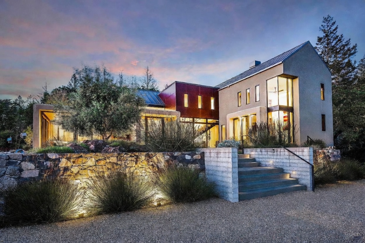 This striking modern residence features a unique blend of architectural styles, combining clean, light-colored stucco walls with a bold, rust-toned metal-clad section. The home is elevated by a rustic stone retaining wall and accessed via a set of concrete stairs, creating a sophisticated tiered landscape. Large floor-to-ceiling windows and warm interior lighting create an inviting, cinematic glow against the twilight sky.