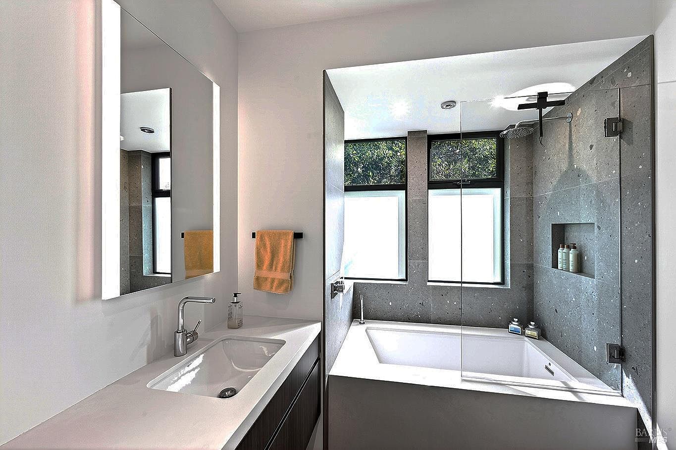 This modern primary bathroom features a sleek vanity with a white countertop and a large, illuminated mirror. Adjacent to the vanity is a luxurious soaking tub enclosed by a glass partition, set against a striking dark stone-tiled wall with a built-in shower niche. The space is bright and airy, with large windows that allow natural light to flood the room, creating a sophisticated and spa-like atmosphere.