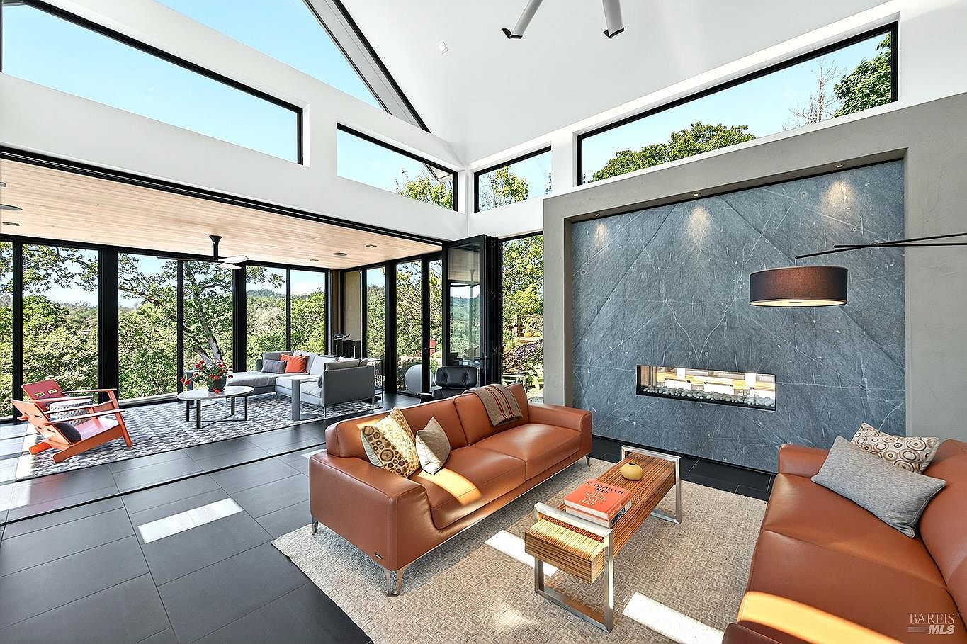This stunning, modern living room features an expansive open-concept design with floor-to-ceiling glass walls that seamlessly blend the indoor space with the surrounding forest. A striking floor-to-ceiling gray stone fireplace serves as the focal point, complemented by rich leather sofas and a minimalist coffee table. The high, vaulted ceilings and clerestory windows flood the room with natural light, creating an airy and sophisticated atmosphere.
