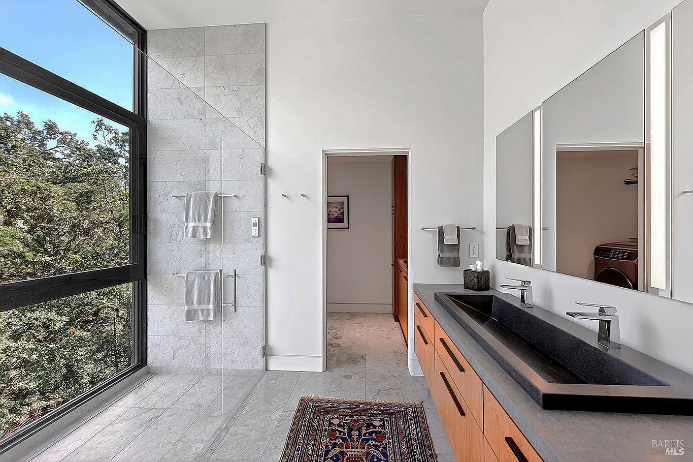 This modern primary bathroom features a sleek, long black trough sink set into a warm wood vanity, complemented by large mirrors and minimalist fixtures. A glass-enclosed shower with light grey stone tile walls stands adjacent to a floor-to-ceiling window that offers a serene view of the surrounding trees. The space is tied together by a traditional patterned rug on the stone floor, creating a sophisticated blend of contemporary design and natural warmth.