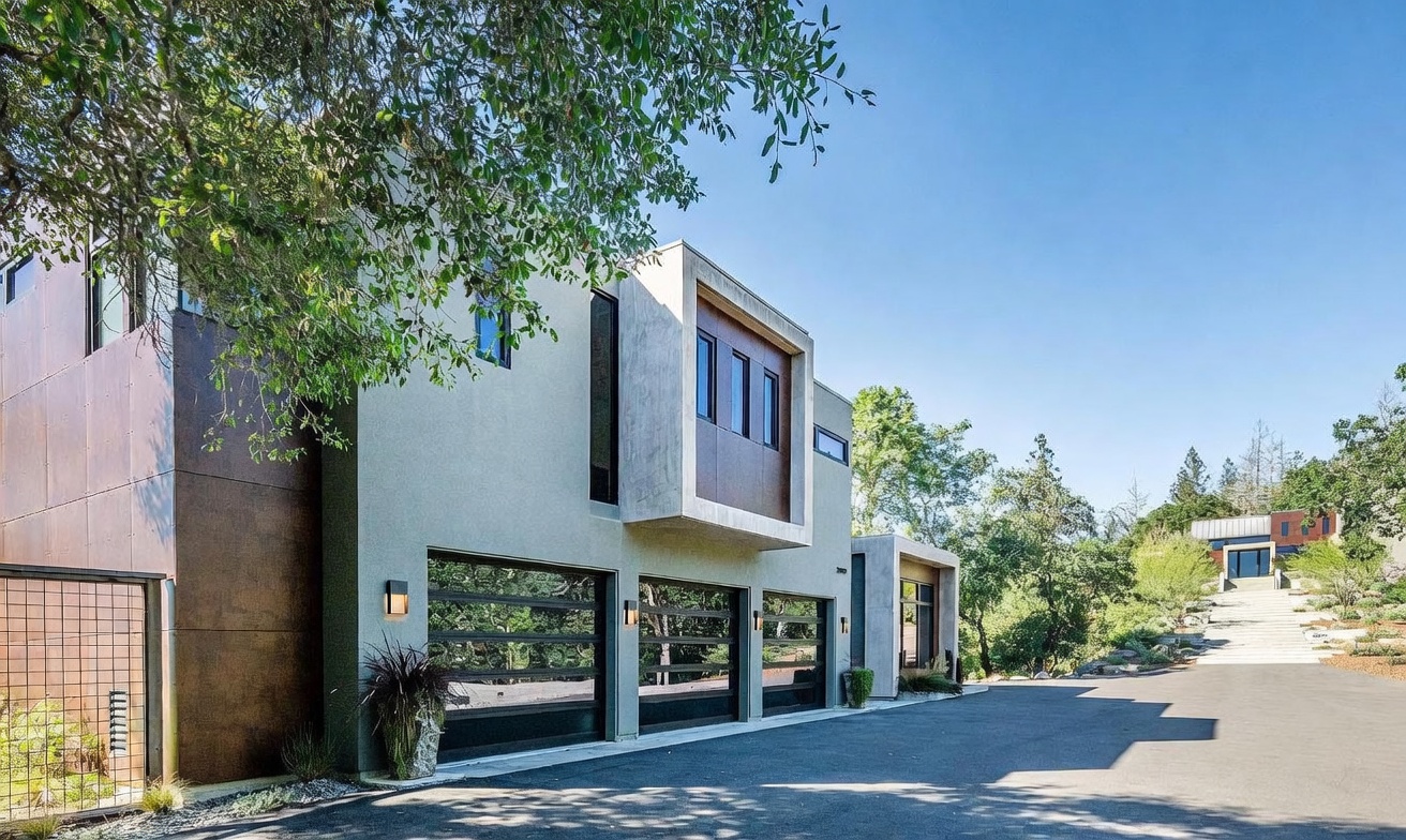 This modern architectural home features a striking three-car garage with sleek, horizontal-paneled glass doors and a contemporary concrete and metal facade. The driveway leads up to the structure, which is nestled among mature trees under a clear blue sky, showcasing a sophisticated blend of industrial and minimalist design. The perspective captures the expansive driveway and the home's unique geometric profile, emphasizing its luxury and curb appeal.