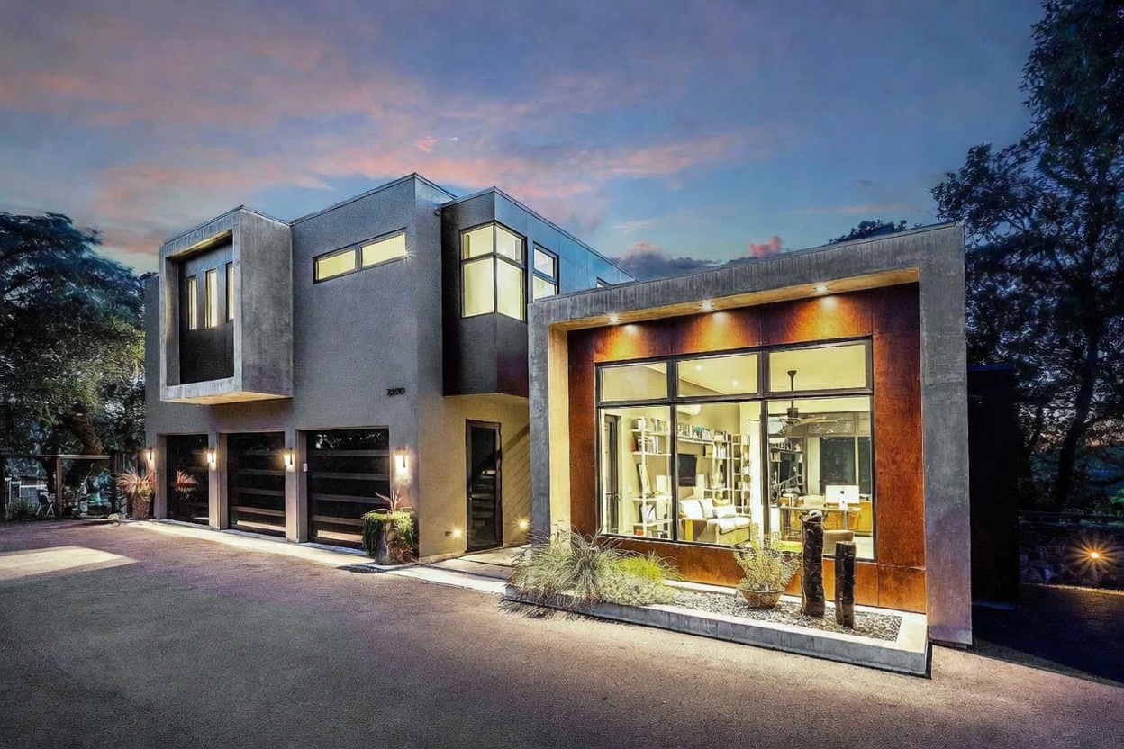 This striking modern architectural home features a bold combination of smooth gray concrete and warm, rusted metal paneling. The front facade includes a multi-car garage on the left and a large, floor-to-ceiling glass-walled office or living space on the right, illuminated by warm interior lighting. The perspective is a low-angle, wide shot that captures the dramatic contrast between the twilight sky and the home's sophisticated, industrial-inspired design.