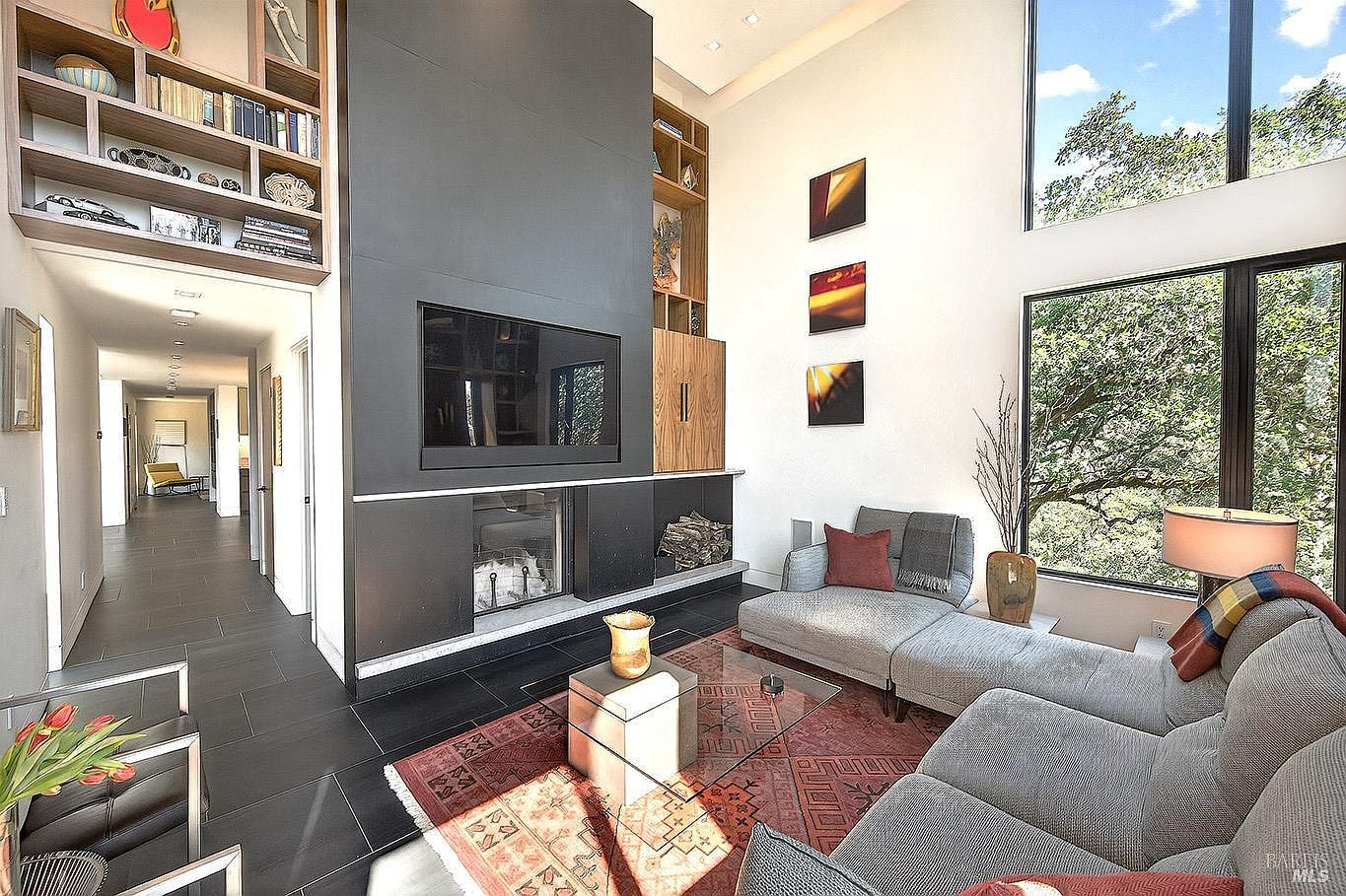 This modern, high-ceilinged living room features a striking floor-to-ceiling dark gray fireplace surround with an integrated television and built-in shelving. A plush gray sectional sofa sits atop a vibrant, patterned area rug, while large windows provide an expansive view of the surrounding trees. The space is characterized by its contemporary design, clean lines, and an open, airy atmosphere that flows into a long hallway.