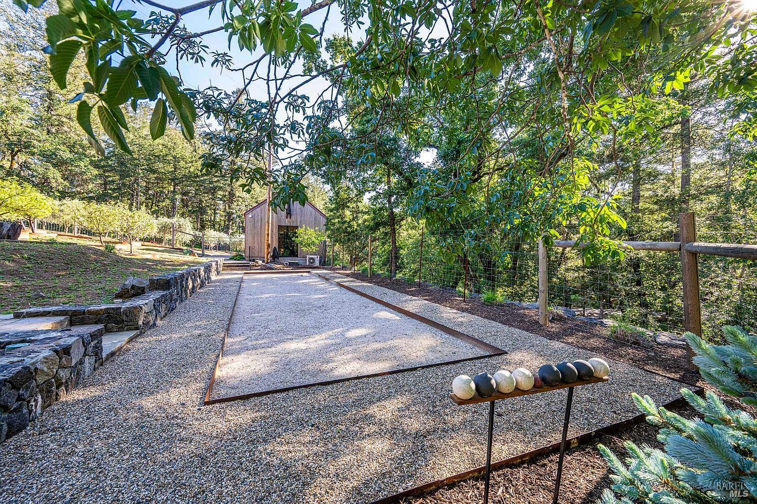 This outdoor space features a well-maintained bocce ball court set against a backdrop of lush, mature trees and a rustic wooden structure. The scene includes a stone retaining wall, a gravel ground cover, and a rack of bocce balls in the foreground, creating an inviting atmosphere for leisure and recreation. The perspective is a wide-angle shot from ground level, emphasizing the serene and natural setting of the property.