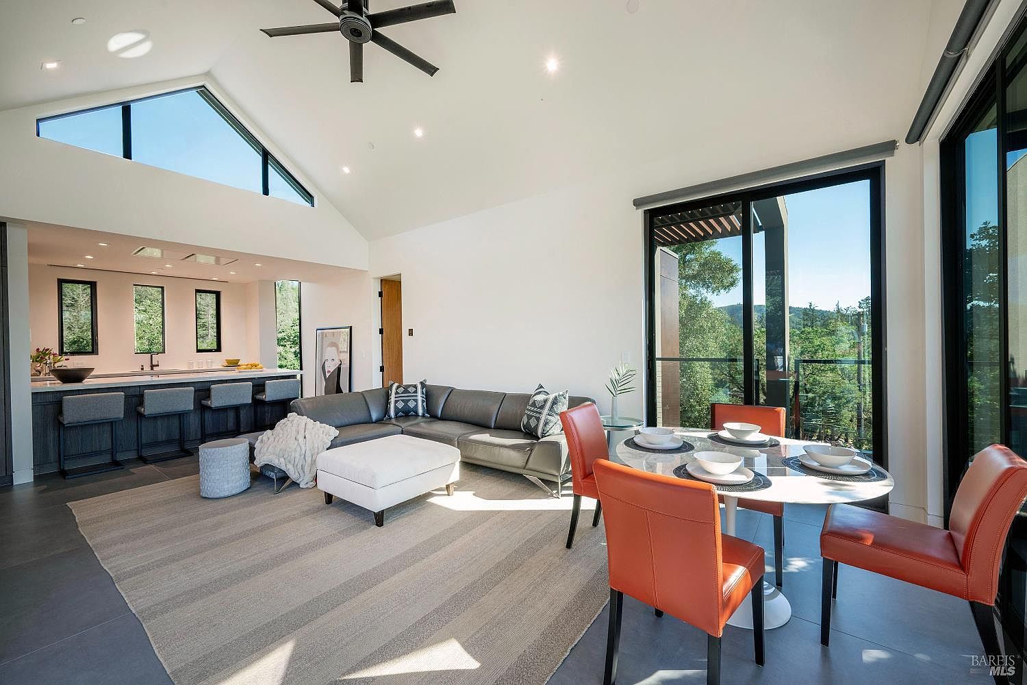 This bright and airy open-concept living space features a vaulted ceiling with a modern ceiling fan and large windows that frame a scenic outdoor view. The room is anchored by a sleek grey sectional sofa, a white ottoman, and a dining area with vibrant orange chairs, creating a sophisticated yet inviting atmosphere. The seamless transition between the living area and the adjacent kitchen, complete with bar seating, highlights the home's contemporary design and spacious layout.