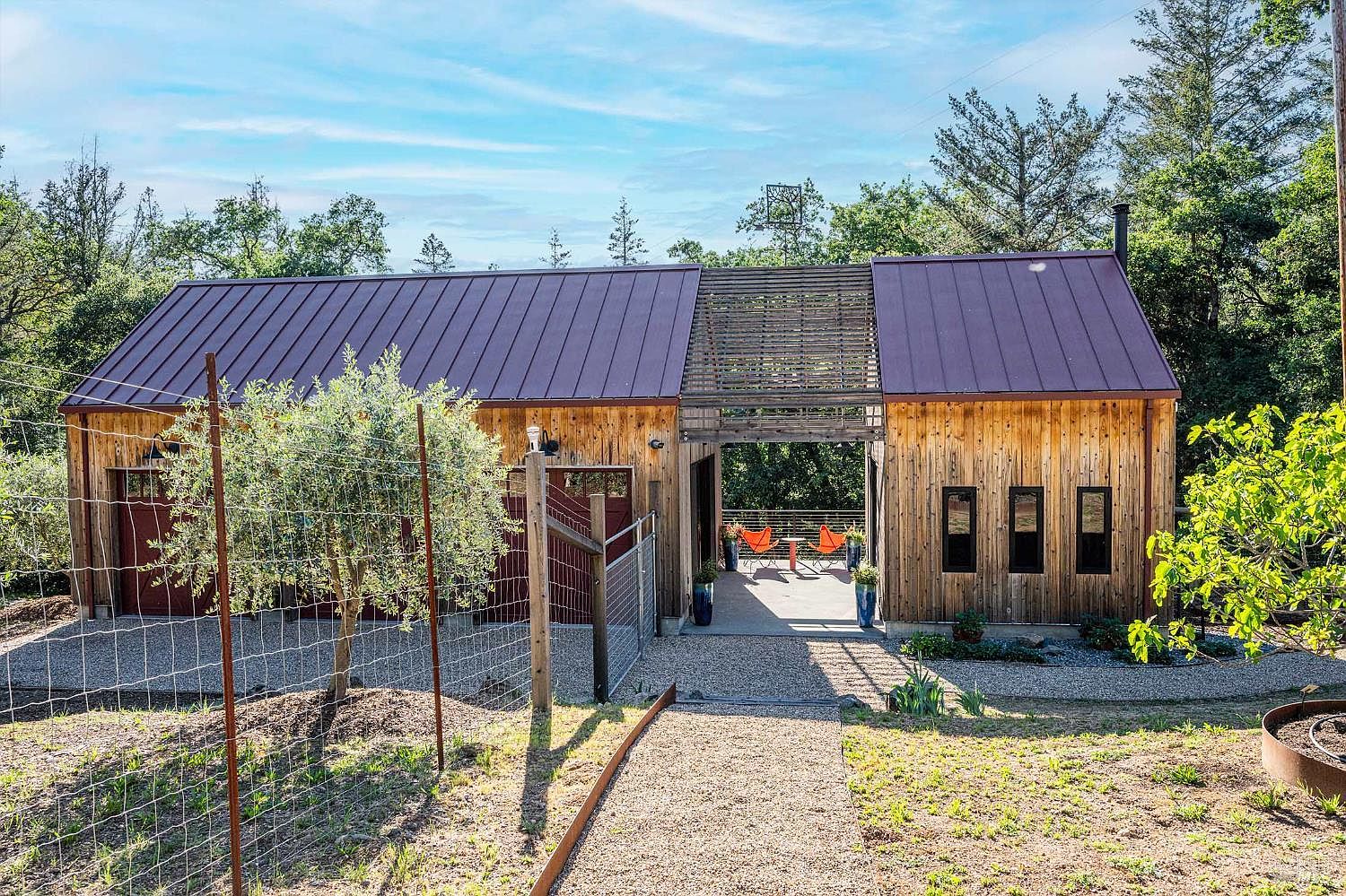 This unique, modern rustic structure features a split-design with two wood-clad buildings connected by a central open-air breezeway topped with a slatted wooden pergola. The exterior showcases warm, vertical wood siding, a dark metal roof, and a gravel courtyard, creating a serene and contemporary aesthetic. The scene is framed by lush greenery and a gravel path, emphasizing a peaceful, nature-integrated living space.