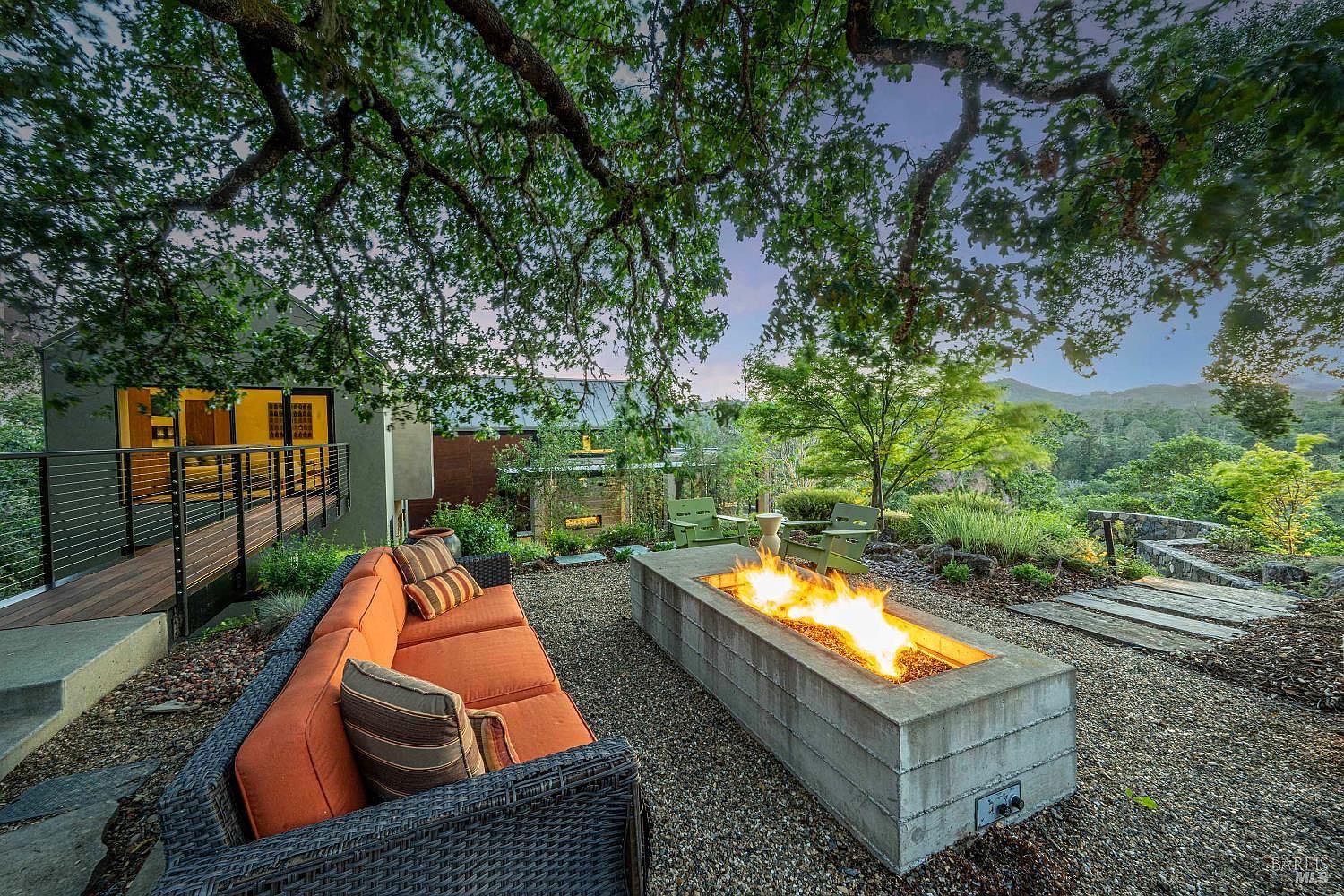 This inviting outdoor living space features a modern concrete fire pit as the centerpiece, surrounded by comfortable orange-cushioned wicker seating. A wooden walkway leads to a contemporary home structure, all set beneath the sprawling canopy of a large oak tree with a scenic, elevated view of the surrounding hills. The atmosphere is serene and sophisticated, perfect for evening relaxation or entertaining.