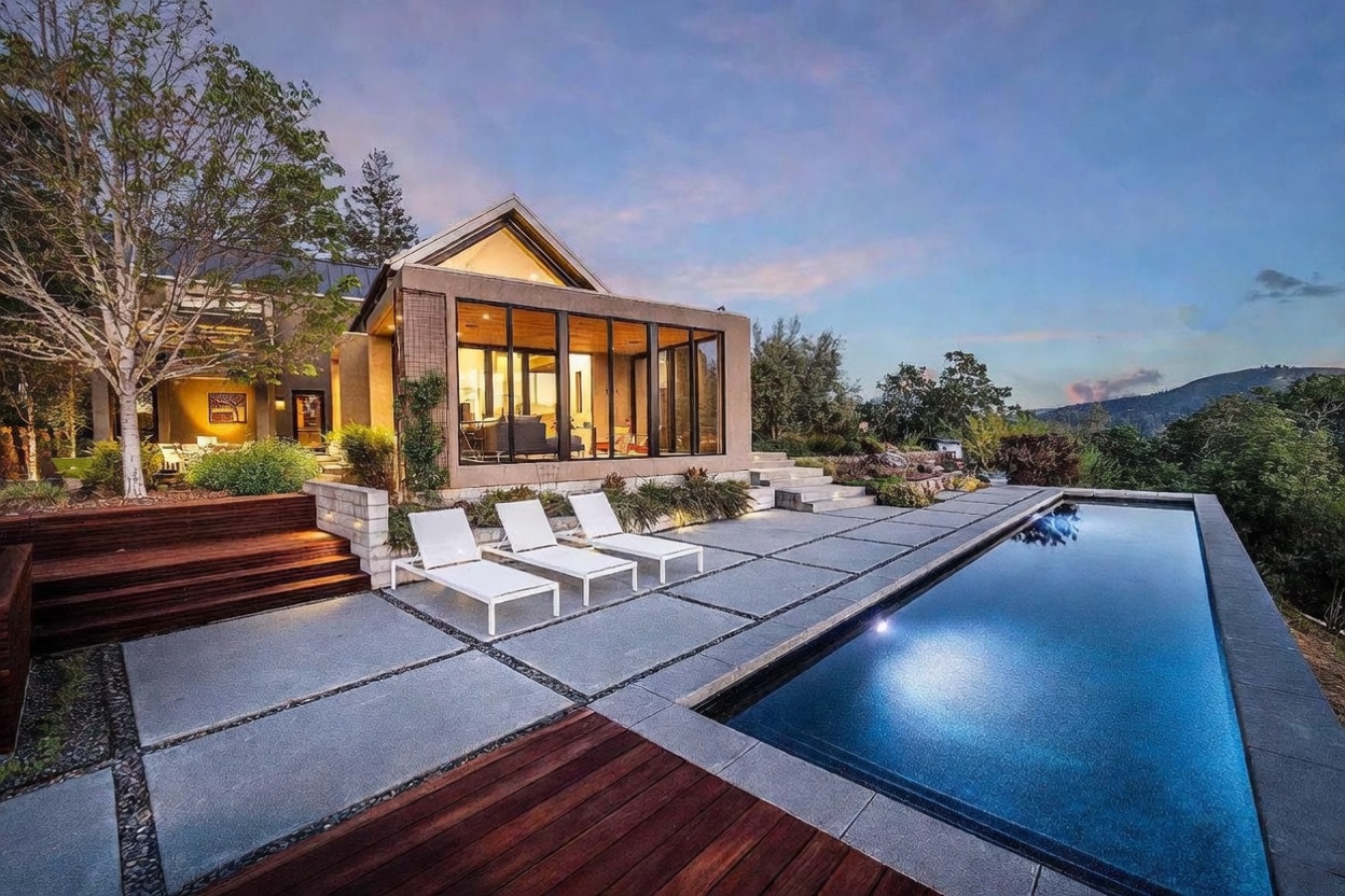 This stunning contemporary residence features a sleek, rectangular lap pool set against a backdrop of rolling hills and a twilight sky. The outdoor living space includes a modern concrete patio with three white lounge chairs, a wooden deck transition, and a glass-walled sunroom that seamlessly blends indoor and outdoor living. The architectural design emphasizes clean lines, natural materials, and an inviting, luxurious atmosphere perfect for evening relaxation.