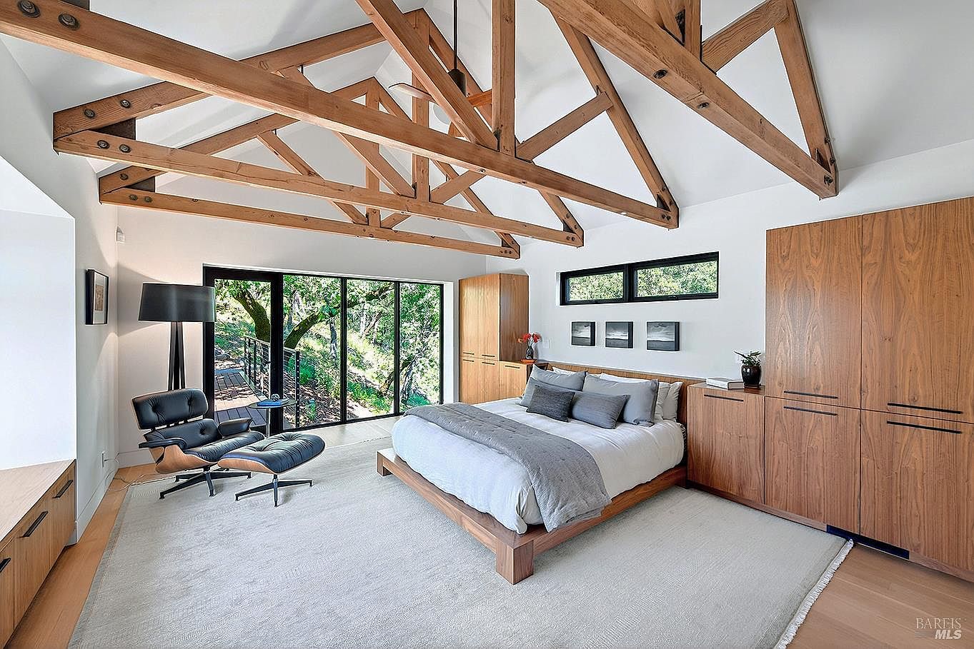 This spacious primary bedroom features a striking vaulted ceiling with exposed wooden trusses, creating a warm, rustic-modern aesthetic. The room is anchored by a low-profile wooden platform bed and complemented by custom built-in cabinetry, a sleek Eames-style lounge chair, and large floor-to-ceiling windows that invite natural light and views of the surrounding forest. The neutral color palette and clean lines contribute to a serene, high-end retreat atmosphere.