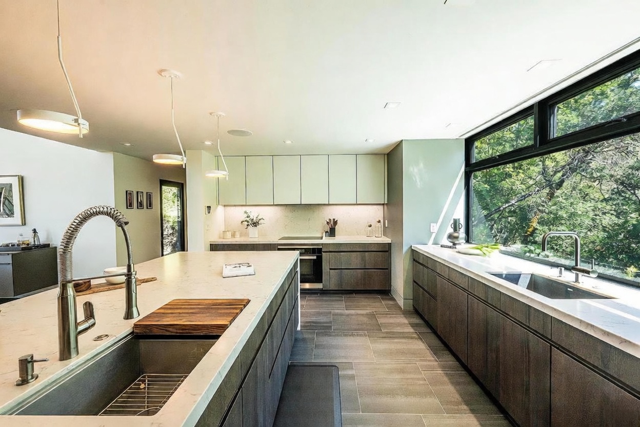 This modern, high-end kitchen features a large central island with a waterfall quartz countertop and a sleek, dark wood finish. The space is illuminated by minimalist pendant lighting and large windows that provide a scenic view of the surrounding greenery. The design combines contemporary aesthetics with functional elements, including integrated appliances and a spacious layout perfect for culinary activities.