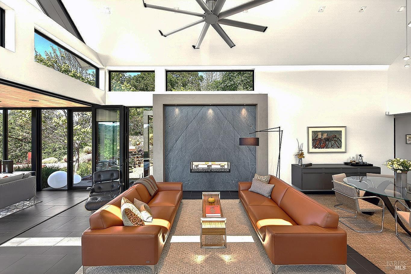 This sophisticated, open-concept living room features a striking floor-to-ceiling dark stone fireplace as its central focal point, complemented by two sleek, cognac-colored leather sofas. The space is bathed in natural light from expansive windows and glass doors that seamlessly connect the interior to the outdoor landscape. A large, modern ceiling fan hangs from the vaulted ceiling, enhancing the airy, contemporary aesthetic of this high-end living area.
