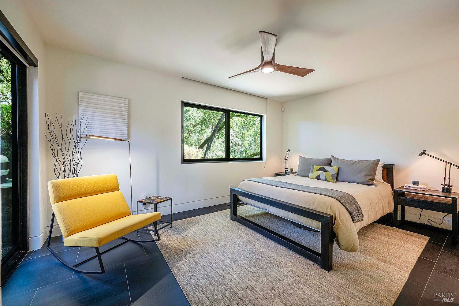 This modern primary bedroom features a minimalist aesthetic with a sleek black-framed bed, a vibrant yellow rocking chair, and dark tile flooring. A large window provides a serene view of the surrounding trees, while a contemporary ceiling fan and floor lamp add functional style to the space. The room is anchored by a large, neutral-toned area rug, creating a calm and sophisticated atmosphere.