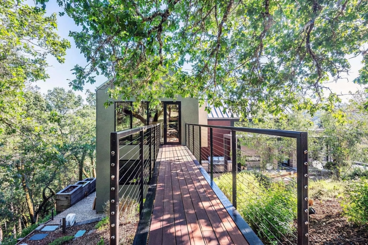 This image captures a modern, elevated wooden walkway leading to the entrance of a contemporary home nestled among mature oak trees. The architectural design features clean lines and a minimalist aesthetic, with a cable-railing system that maintains an open, airy feel while connecting the structure to the surrounding natural landscape. The perspective is a straight-on, eye-level shot that draws the viewer toward the glass-paned door, emphasizing the seamless integration between the modern residence and the wooded environment.